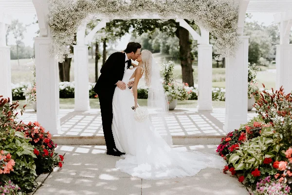 A bride and groom kissing under a white floral arch with surrounding flower arrangements.