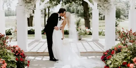 A bride and groom kissing under a white floral arch with surrounding flower arrangements.