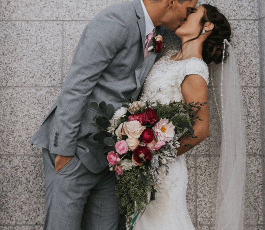 A bride and groom kissing while the bride holds a mixed floral bouquet and the groom wears a matching boutonniere.
