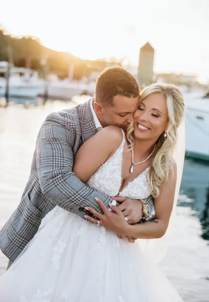 A bride with styled hair and makeup embracing a groom in a waterfront setting.