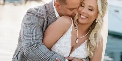 A bride with styled hair and makeup embracing a groom in a waterfront setting.