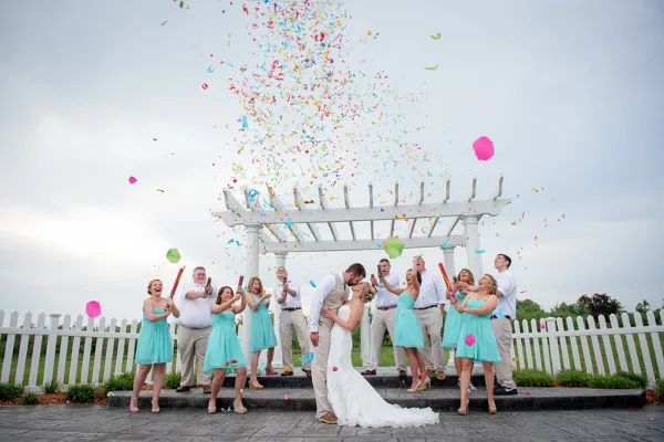 A bride and groom kiss under a white gazebo while their wedding party throws colorful confetti.
