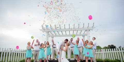 A bride and groom kiss under a white gazebo while their wedding party throws colorful confetti.