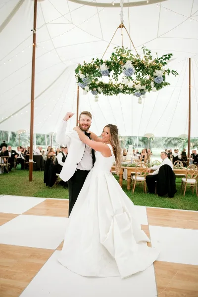 A bride and groom dancing at a wedding reception under a floral chandelier in a white tent.