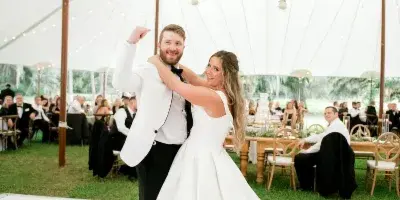 A bride and groom dancing at a wedding reception under a floral chandelier in a white tent.