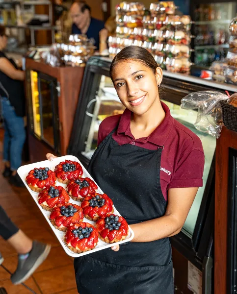 A smiling bakery employee in a maroon shirt and black apron holding a tray of fresh fruit tarts in a bakery setting.