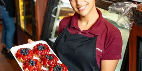 A smiling bakery employee in a maroon shirt and black apron holding a tray of fresh fruit tarts in a bakery setting.