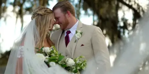 A bride and groom sharing a tender moment outdoors, with the bride holding a white bouquet and the groom wearing a beige suit.
