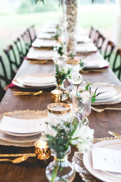 Elegant outdoor wedding reception table set with gold-rimmed plates, glassware, candles, and floral centerpieces.