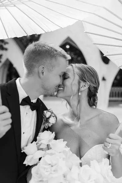 A black and white photograph of a bride and groom smiling at each other under a white parasol, with the bride holding a bouquet of white roses.
