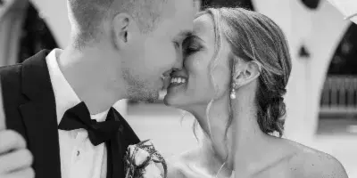 A black and white photograph of a bride and groom smiling at each other under a white parasol, with the bride holding a bouquet of white roses.