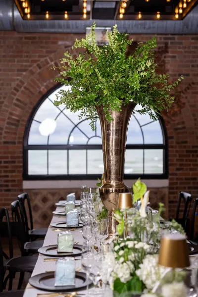 A long event table set with a tall green floral arrangement in a bronze vase, white flowers, and place settings in a brick-walled venue.