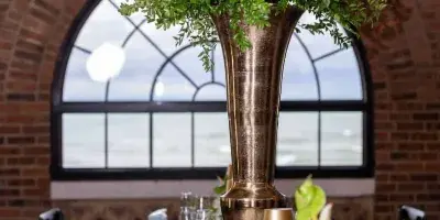 A long event table set with a tall green floral arrangement in a bronze vase, white flowers, and place settings in a brick-walled venue.