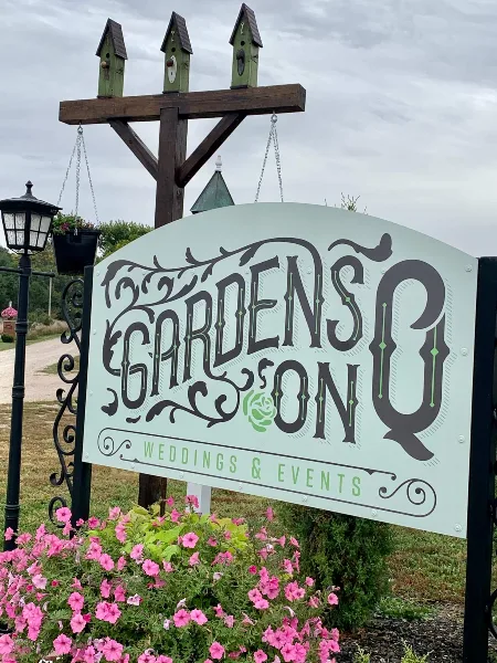 A decorative entrance sign for Gardens on Q Weddings & Events featuring ornate typography, birdhouses, and pink flowers.