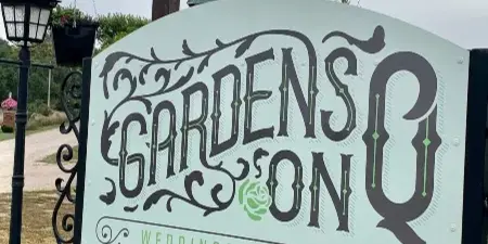 A decorative entrance sign for Gardens on Q Weddings & Events featuring ornate typography, birdhouses, and pink flowers.
