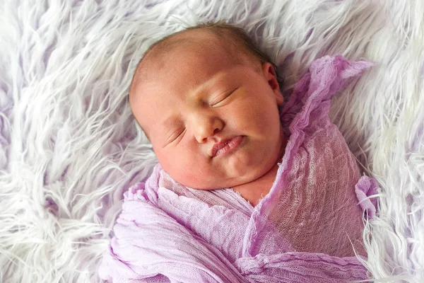 A sleeping newborn baby wrapped in a purple swaddle on a white fluffy blanket.