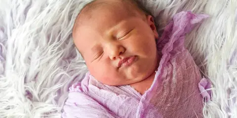 A sleeping newborn baby wrapped in a purple swaddle on a white fluffy blanket.