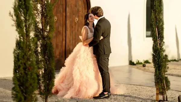 A bride in a pink gown and a groom in a black suit embracing in front of a white building with large wooden doors and tall green topiaries.