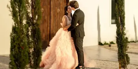 A bride in a pink gown and a groom in a black suit embracing in front of a white building with large wooden doors and tall green topiaries.