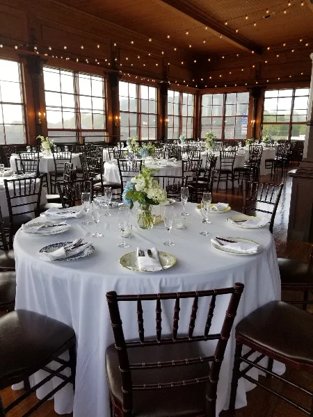 Interior view of a historic wooden hall with large windows, string lights, and round tables set with white linens and floral centerpieces for a wedding reception.