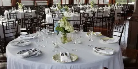 Interior view of a historic wooden hall with large windows, string lights, and round tables set with white linens and floral centerpieces for a wedding reception.