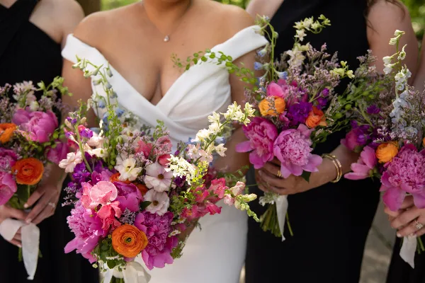 A bride and bridesmaids holding colorful wedding bouquets featuring pink peonies, orange ranunculus, and white snapdragons.