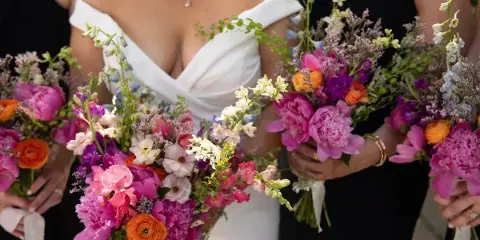 A bride and bridesmaids holding colorful wedding bouquets featuring pink peonies, orange ranunculus, and white snapdragons.