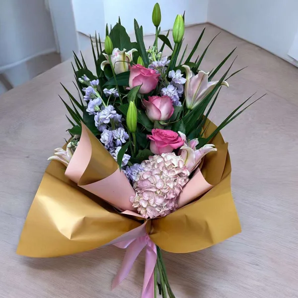 A floral bouquet containing pink roses, white lilies, and pale hydrangeas wrapped in gold and pink paper.