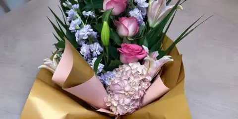 A floral bouquet containing pink roses, white lilies, and pale hydrangeas wrapped in gold and pink paper.