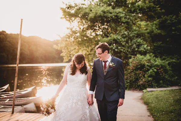 A bride and groom walking hand-in-hand along a path near a lake during a wedding photoshoot.