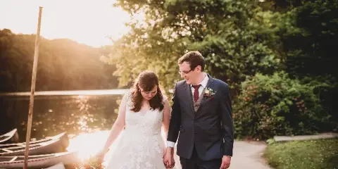 A bride and groom walking hand-in-hand along a path near a lake during a wedding photoshoot.