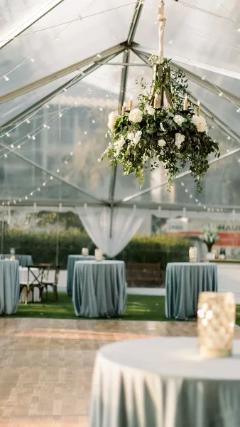 A clear-frame event tent interior featuring a hanging floral chandelier, string lights, and round tables with grey tablecloths.