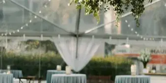 A clear-frame event tent interior featuring a hanging floral chandelier, string lights, and round tables with grey tablecloths.