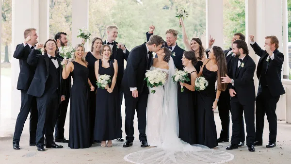 A wedding party including the bride and groom posing together outdoors under a colonnade.