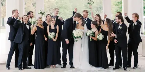 A wedding party including the bride and groom posing together outdoors under a colonnade.