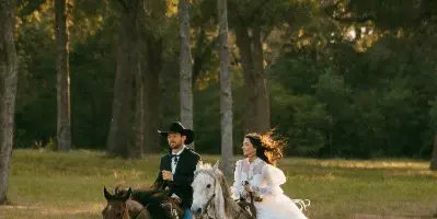 A bride and groom riding horses in a grassy field with trees in the background.