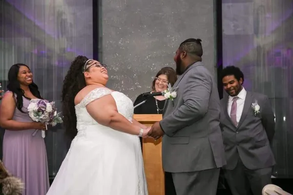 A bride and groom holding hands at a podium during a wedding ceremony with an officiant and wedding party members in the background.