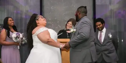 A bride and groom holding hands at a podium during a wedding ceremony with an officiant and wedding party members in the background.