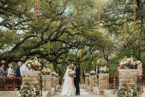 A bride and groom sharing a kiss during an outdoor wedding ceremony under a large tree with hanging lights and floral arrangements.