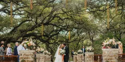 A bride and groom sharing a kiss during an outdoor wedding ceremony under a large tree with hanging lights and floral arrangements.
