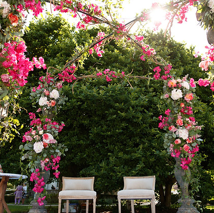 A floral wedding arch decorated with pink and white flowers and greenery, with two white chairs set up in front of a hedge.