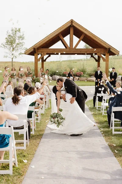 A bride and groom share a kiss at a wooden outdoor arbor during a wedding ceremony, with guests seated in white chairs on either side of a paved walkway.