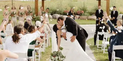 A bride and groom share a kiss at a wooden outdoor arbor during a wedding ceremony, with guests seated in white chairs on either side of a paved walkway.