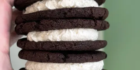 A hand holding a stack of chocolate cookies with white buttercream filling.