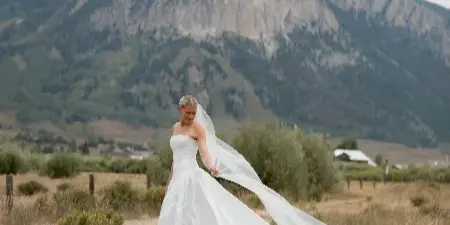 A bride in a white wedding dress and veil standing in a grassy field with a large mountain peak in the background.