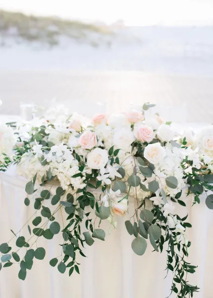 A wedding table runner featuring white and blush roses, hydrangeas, and trailing eucalyptus on a white tablecloth.