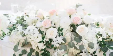 A wedding table runner featuring white and blush roses, hydrangeas, and trailing eucalyptus on a white tablecloth.