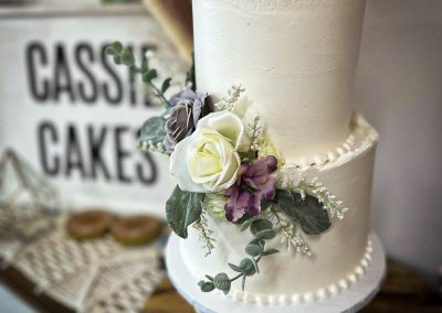 A white two-tier wedding cake decorated with white and purple flowers, displayed in front of a Cassie Cakes sign.