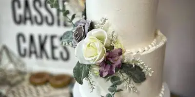 A white two-tier wedding cake decorated with white and purple flowers, displayed in front of a Cassie Cakes sign.