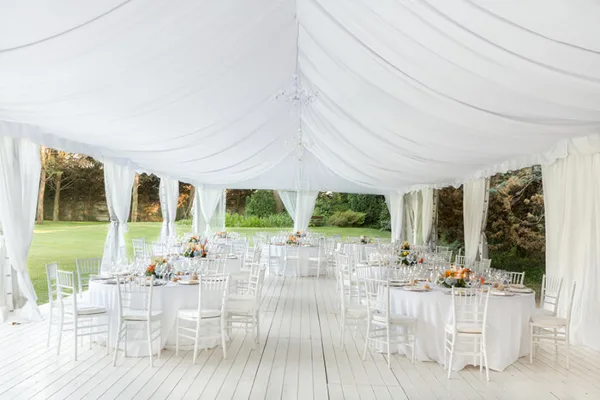 A large white event tent with draped fabric, set up with round tables and chairs for a wedding or party.